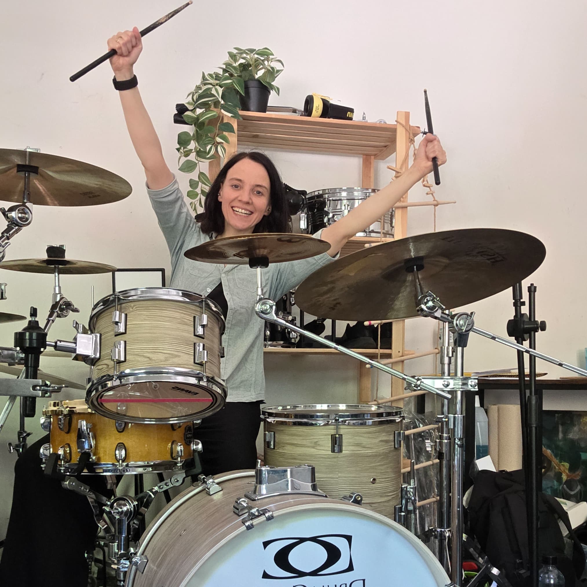 Eine freudige Schlagzeugerin sitzt hinter ihrem Drumset und streckt beide Trommelstöcke triumphierend nach oben.